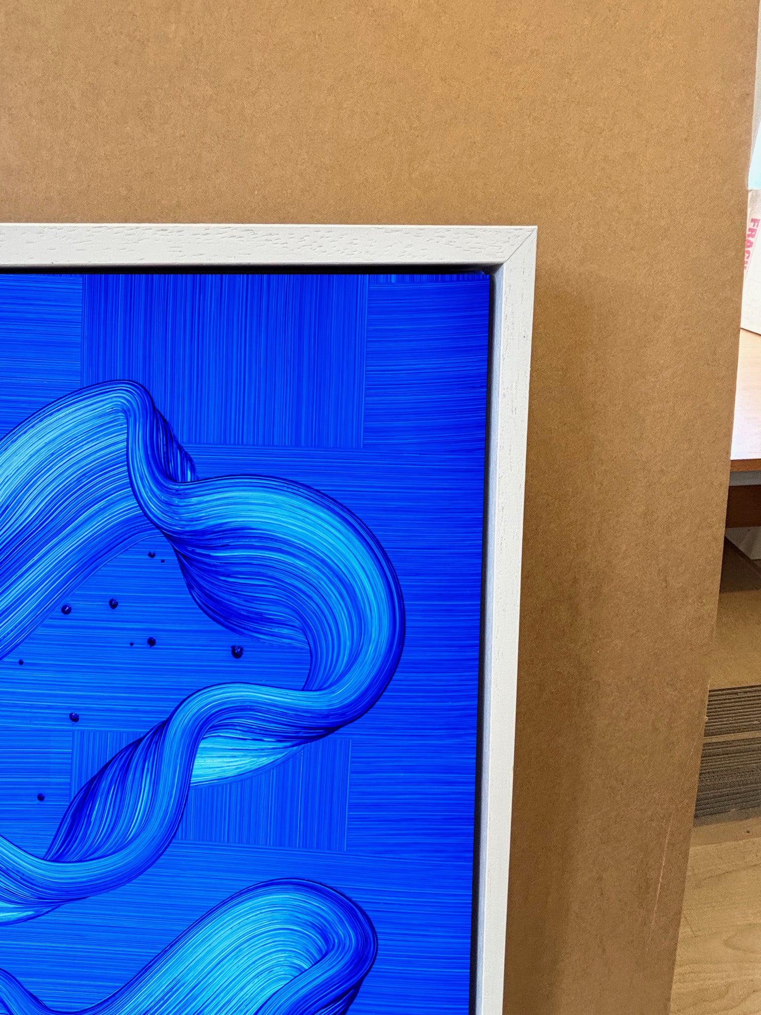 Ebb and Flow abstract art by Lara Sayegh, featuring ocean-inspired rhythmic brushstrokes, printed on canvas stretched in a white floater tray frame on a wooden floor with a brown wall background.