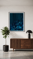 A framed abstract art print featuring deep blue and teal tones, displayed on a light gray wall above a wooden sideboard with a potted plant and a black table lamp.