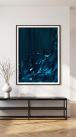 A framed abstract art print featuring deep teal and blue textured brushstrokes, displayed on a white wall above a black console table with a white vase of branches.